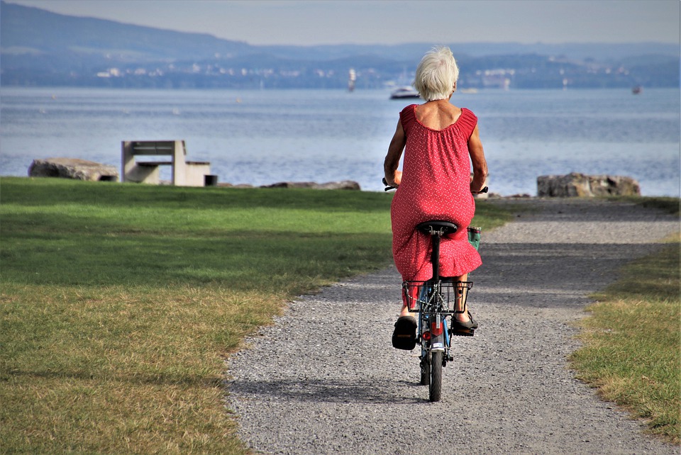 imagem das costas de uma mulher idosa usando um vestido vermelho e andando de bicicleta em estrada próxima a um lago