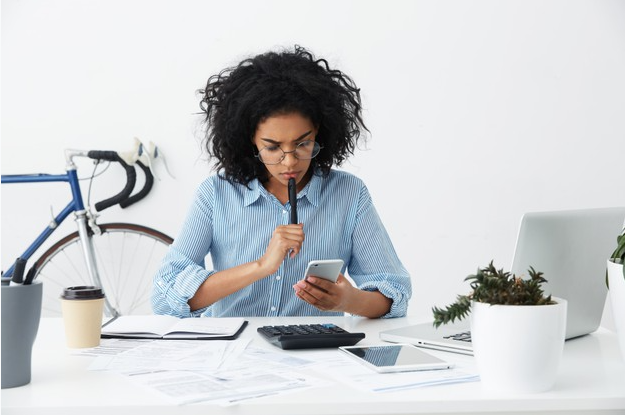 imagem de uma mulher usando camisa azul sentada a uma mesa com um computador olhando para seu celular