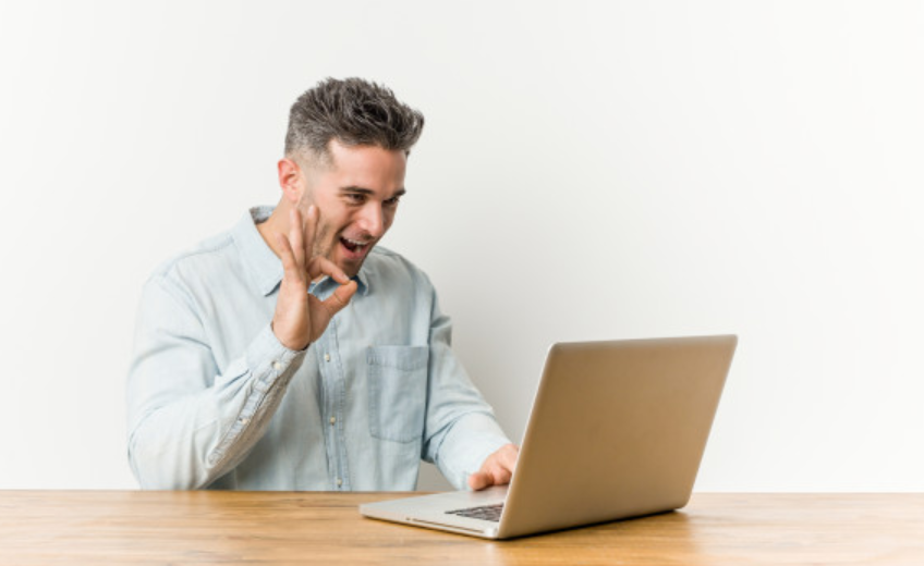 imagem de um homem sentado a uma mesa em frente ao seu laptop fazendo sinal de positivo com a mão