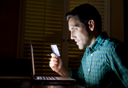 homem usando seu laptop em sala escura segurando seu cartão de crédito com a mão direita