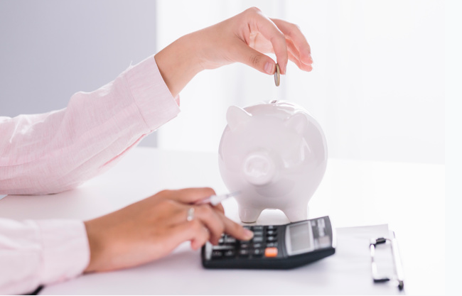 imagem ampliada de mulher usando camisa branca colocando uma moeda dentro de uma cofre branco em formato de porco e usando uma calculadora