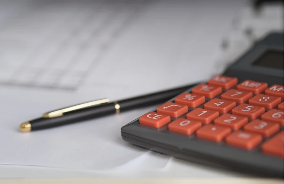 calculadora cinza com botões vermelhos ao lado de uma caneta preta e dourada em cima de uma mesa com papéis