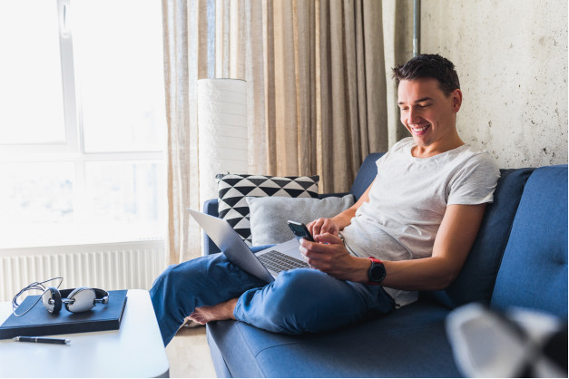 homem usando camisa branca e calça jeans azul sentado em seu sofá sorrindo com um computador no colo enquanto mexe em seu celular