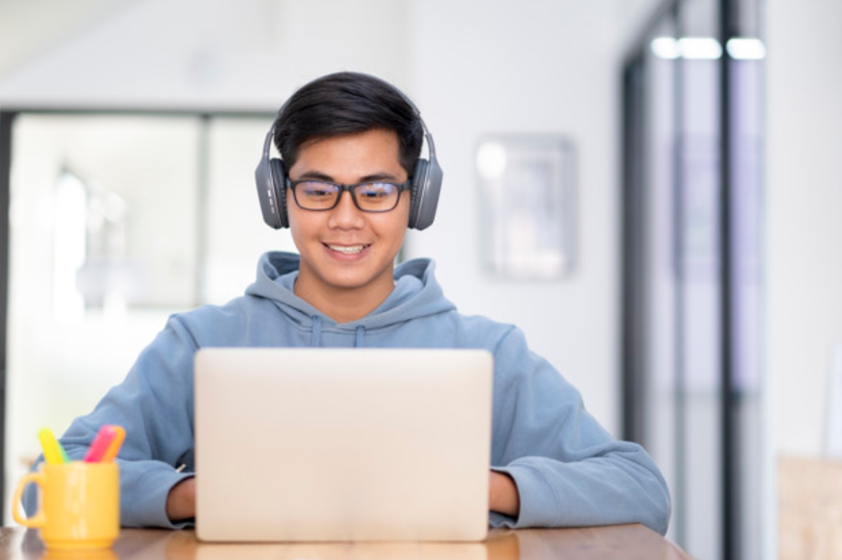 garoto de óculos e moletom azul e usando fones de ouvido, estuda com seu laptop
